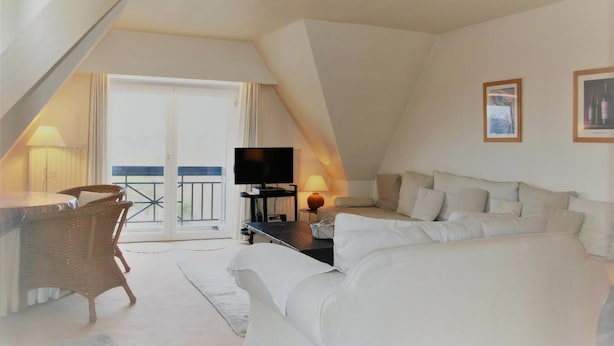 Um quarto estilo loft aconchegante com um sofá branco, uma mesa de jantar, cadeiras de vime, uma TV de tela plana e uma varanda com corrimão.