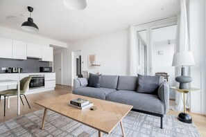 Ein modernes Wohnzimmer mit einer grauen Couch, einem Holztisch und einer Küche im Hintergrund.