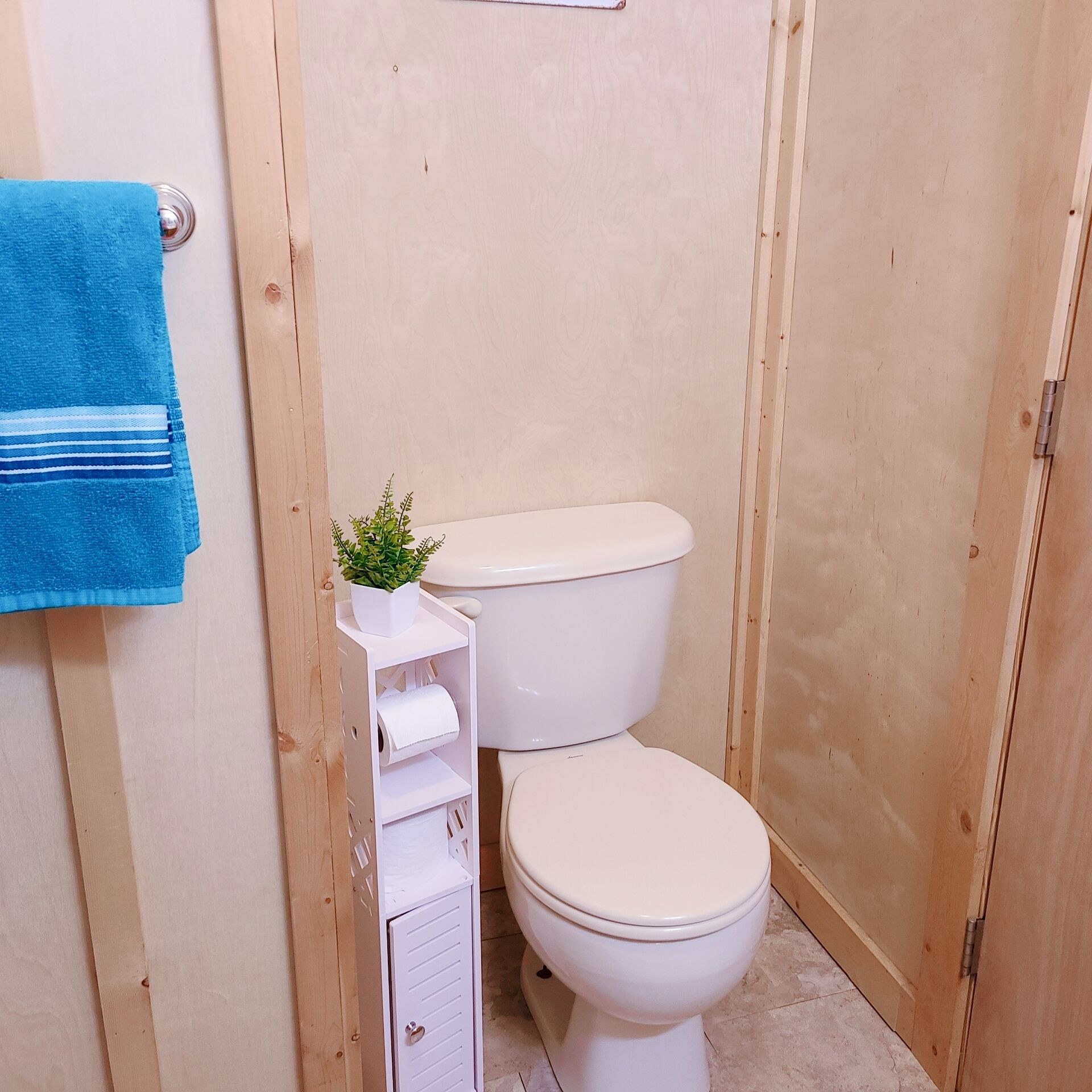 Um banheiro pequeno de madeira com vaso sanitário, prateleira com uma planta e papel higiênico, e uma toalha azul pendurada na parede.