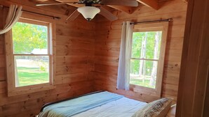 Un dormitorio de cabaña de madera con cama, ventilador de techo y dos ventanas con cortinas.