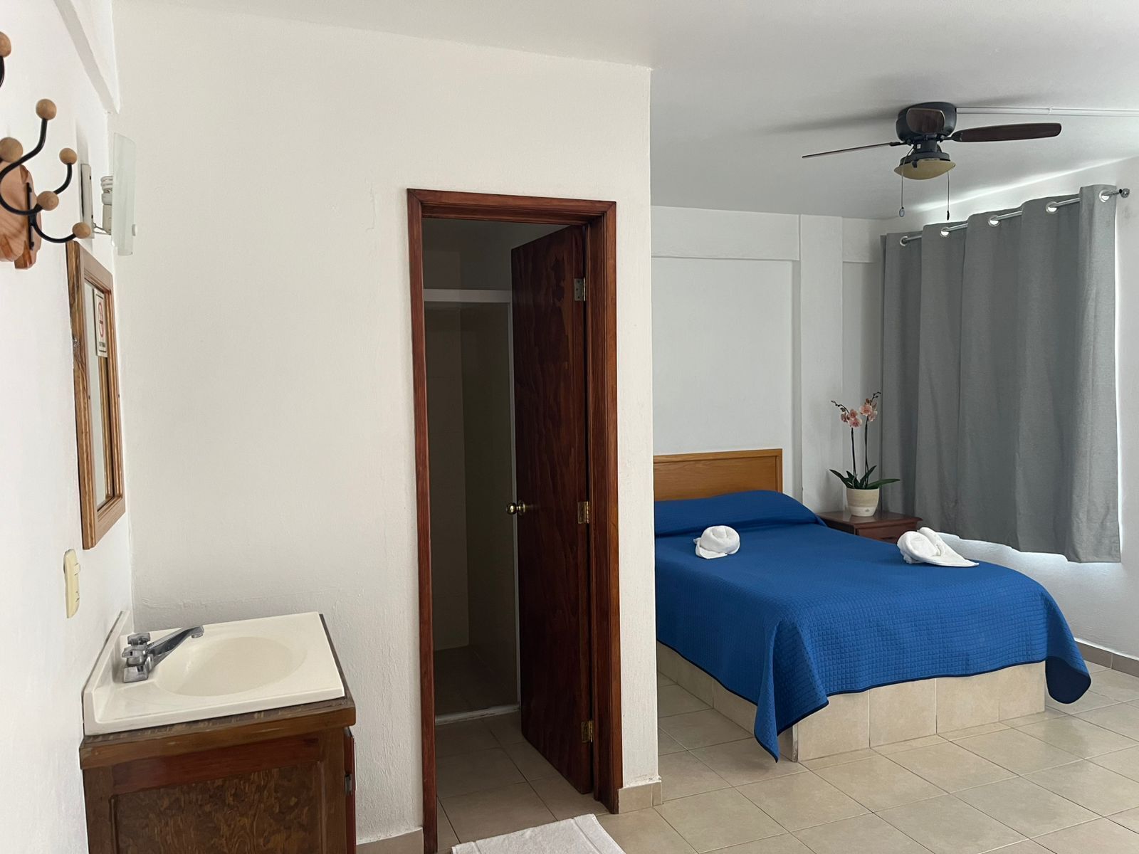 Habitación de hotel con cama, baño con lavamanos, ventilador de techo y ventana con cortinas.