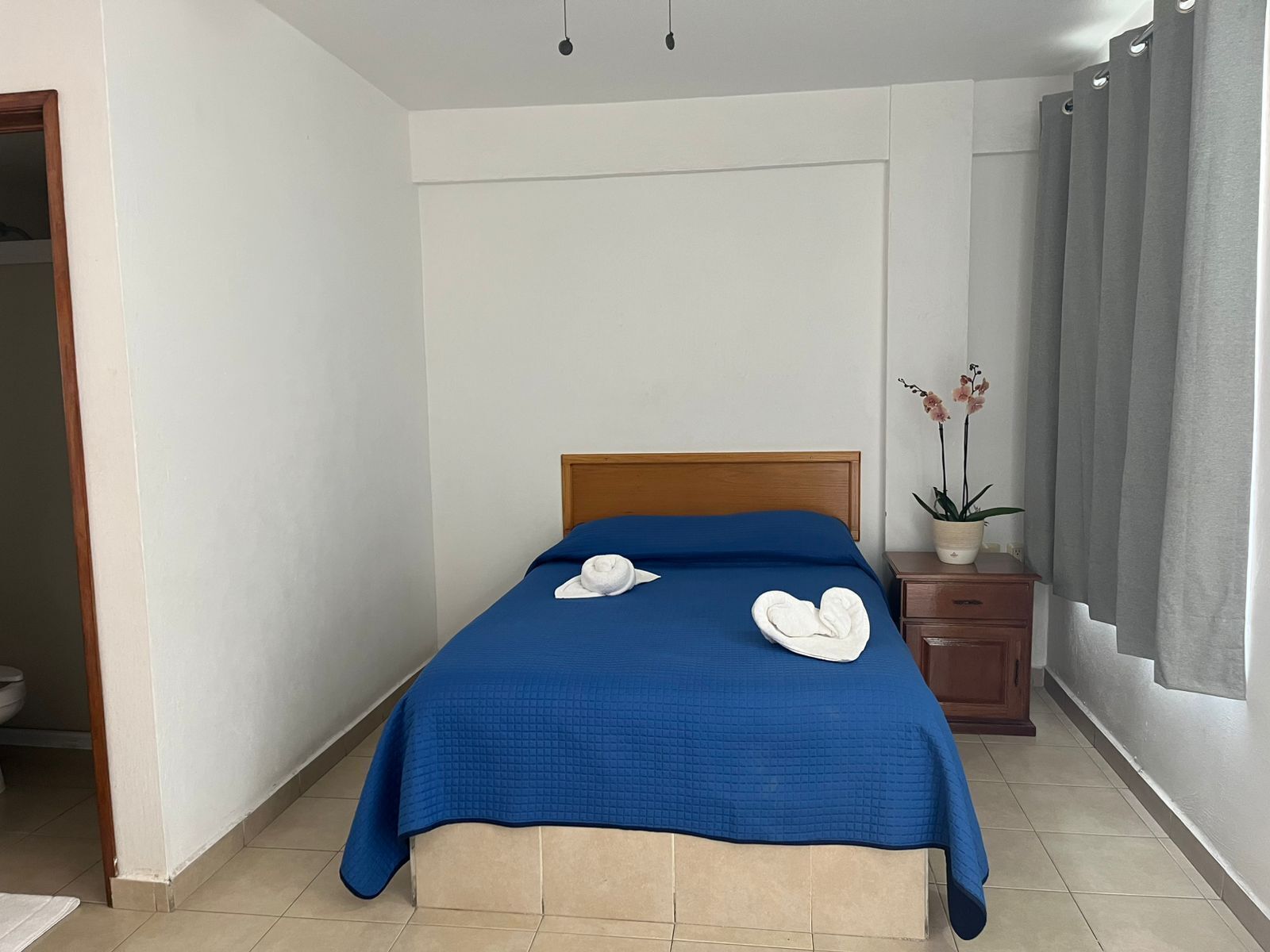 A neatly made bed with a blue bedspread and white towels, a wooden headboard, a small wooden nightstand with a potted orchid, and a grey curtain on the window.