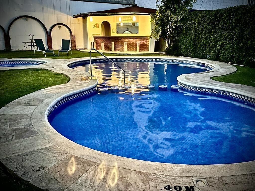 Una piscina con borde de azulejos, un área de barbacoa de ladrillo y una zona de estar cubierta con sillas.
