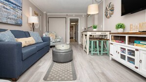 A living room with a blue sofa, a grey ottoman, a coffee table, a white desk with stools, and a TV mounted on the wall. - Oceanfront | Amazing Views & Sunrises (Myrtle Beach)