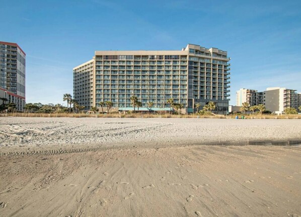 A beachfront hotel with a large building, sandy beach, and palm trees.