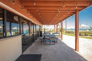 An outdoor patio with wooden tables and benches, a covered seating area with string lights, and a view of the beach.