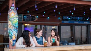 Ein Holztisch mit drei Personen, einer Surfbrettdarstellung und einem Schild für 'Beachfront Kitchen & Bar' mit der Aufschrift 'Musik, Essen, Getränke'.