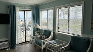 Ein Zimmer mit Blick durch eine Schiebeglastür, eine weiße Klimaanlage, ein petrolfarbener Sessel, ein weißer Korbstuhl, ein kleiner Tisch mit Lampe sowie ein an der Wand befestigter Fernseher.