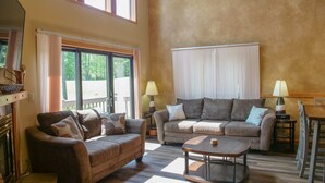 A living room with two sofas, a coffee table, and two lamps. There are large windows with curtains and a view of greenery outside. - Roomy Beautiful Golf/Ski Condo (Clymer)
