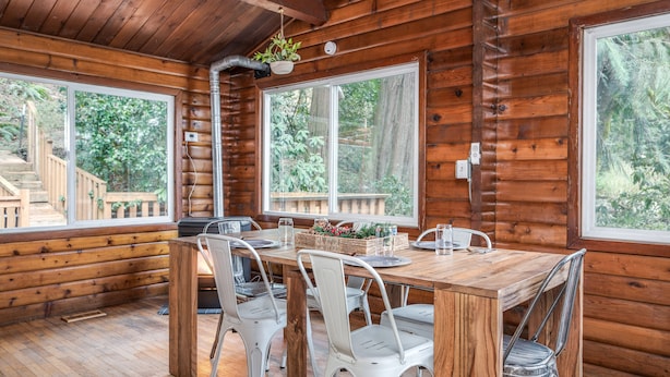 Interno di una baita in legno con un tavolo da pranzo, sedie in metallo bianco e ampie finestre che si affacciano sulla foresta circostante.