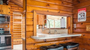 Interno di una baita in legno con angolo cottura, dotato di forno a microonde, forno e lavello. Sono presenti due sgabelli e un cartello appeso al muro.