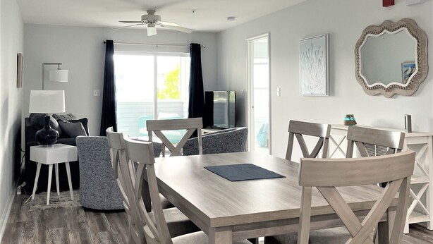 A modern living room with a wooden dining table, comfortable seating, a ceiling fan, and a view of the outdoors. - Coastal Retreat - 2BR CONDO (Rehoboth Beach)