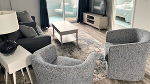 A modern living room with a sofa, armchairs, a coffee table, a TV, and a sliding glass door leading to a balcony. - Coastal Retreat - 2BR CONDO (Rehoboth Beach)