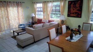 A living room with a sofa, ottoman, dining table, chairs, and a coffee table. There are two lamps on the table and a painting on the wall. - Caye Manje Holiday Lodge (Gros Islet)