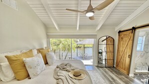 Une chambre à coucher avec un lit, des oreillers, une couverture, un chapeau, un ventilateur de plafond et une vue sur l’extérieur.
