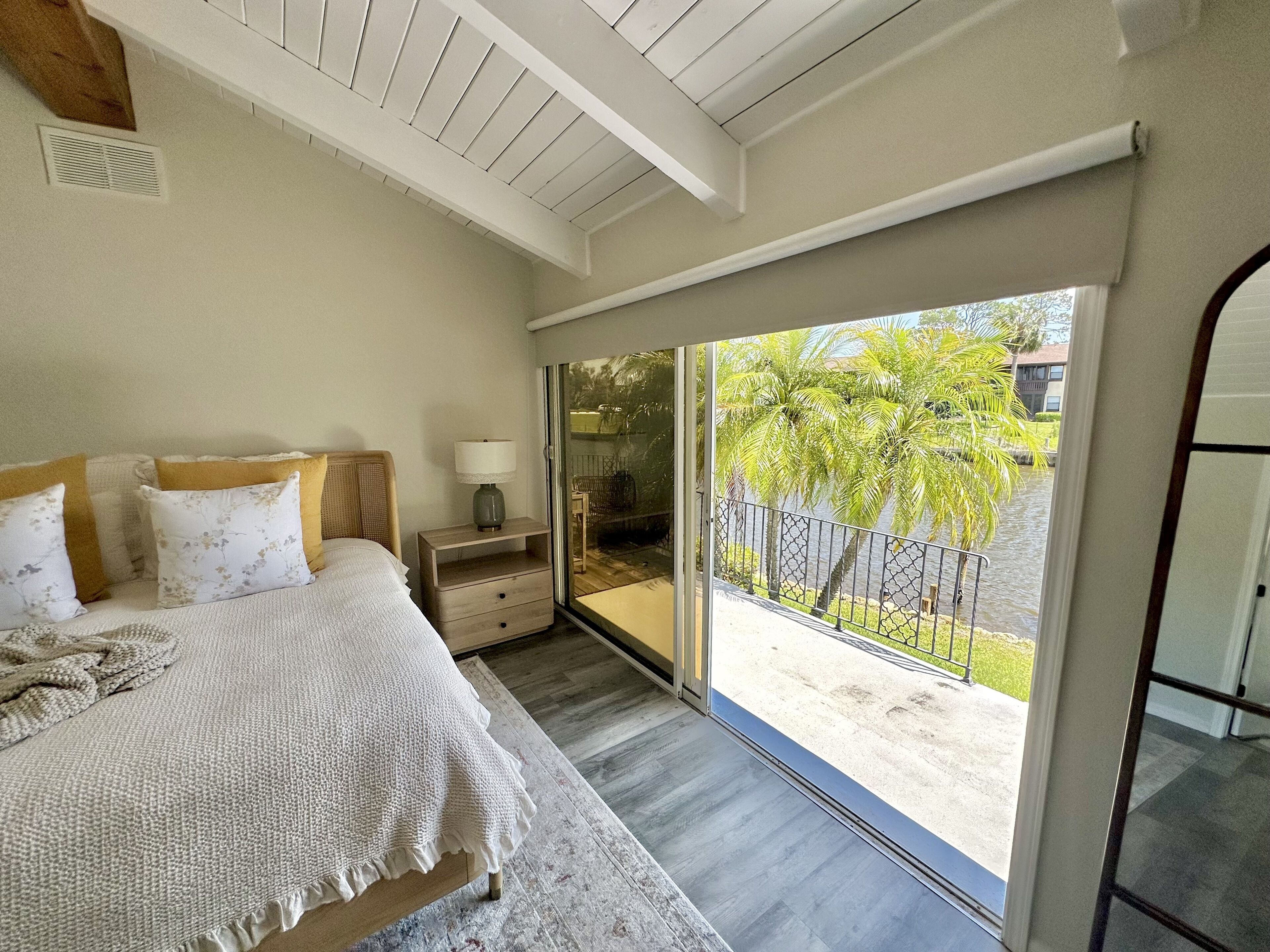 A bedroom with a large window, a bed with a white blanket, a nightstand with a lamp, and a view of palm trees and a body of water.