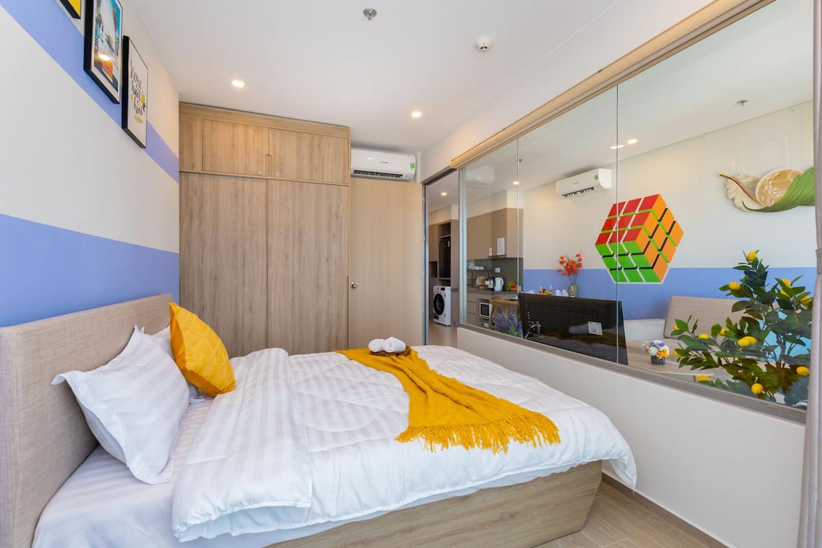Une chambre d’hôtel moderne équipée d’un lit, d’une armoire, d’un climatiseur, d’un bureau avec une chaise et d’une fenêtre donnant sur une plante et un élément décoratif mural.