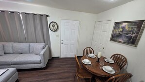 Un salón con un sofá gris, una mesa de comedor con platos, vasos y una vela, y un cuadro en la pared.
