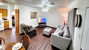 Una sala de estar con sofá, sillón y reposapiés, una televisión y un comedor con mesa dispuesta para dos personas.