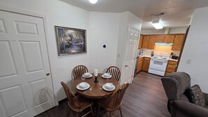 Un comedor con una mesa y sillas de madera, un cuadro en la pared y una cocina al fondo.