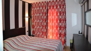 Une chambre d’hôtel avec un lit à rayures, une fenêtre avec des stores, un bureau avec une chaise et un climatiseur fixé au mur.