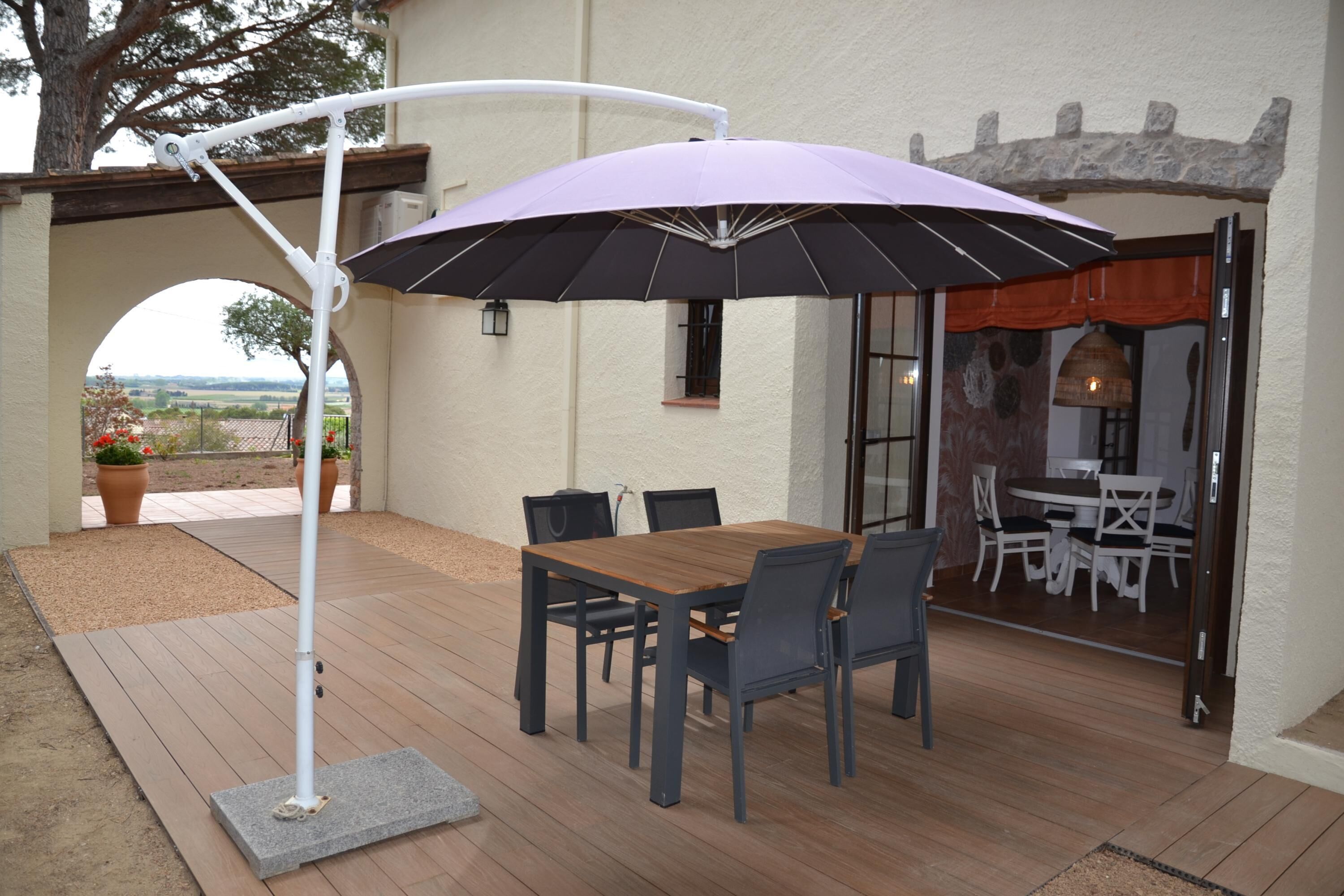 Eine Terrasse im Freien mit einem Holztisch und -stühlen, einem violetten Sonnenschirm und einem weißen Laternenpfosten.