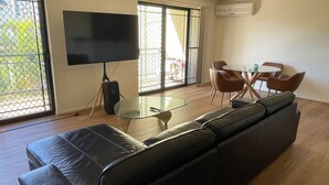 A living room with a black leather sofa, a glass coffee table, a flat-screen TV on a stand, and a dining area with a small round table and four chairs. - Nearby Showgrounds and James St and King Williams Precints (Fortitude Valley)