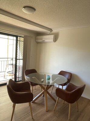 Una habitación con una mesa de vidrio rodeada por cuatro sillas marrones, una lámpara en el techo y un equipo de aire acondicionado.