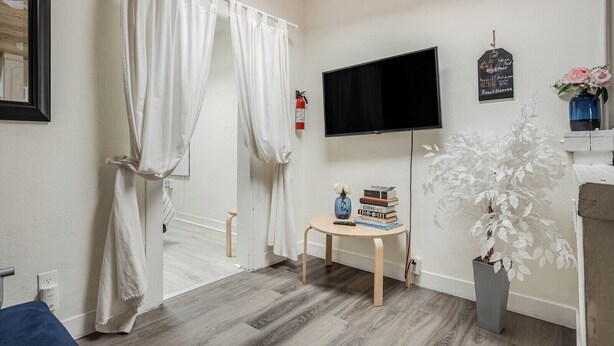 A room with a wooden floor, a bed with white curtains, a television mounted on the wall, a small table with books and a vase, and a potted plant. - Beach Bungalow, Free Parking, 2 blocks to the beach (San Diego)