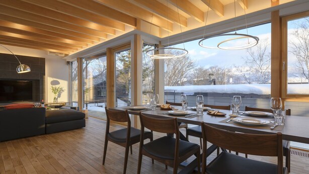 Ein moderner Essbereich mit einem Holztisch, gedeckt für vier Personen, große Fenster mit Blick auf eine verschneite Landschaft und eine gemütliche Sitzecke mit einem Flachbildfernseher.