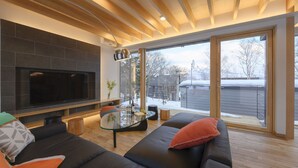 Ein modernes Wohnzimmer mit großem Flachbildfernseher, Glastisch und einer Couch mit Kissen.