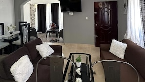 A living room with two sofas, a coffee table, a TV mounted on the wall, and a dining area in the background.
