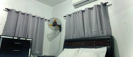 Un dormitorio con cama, aire acondicionado, ventilador de pared y cortinas.