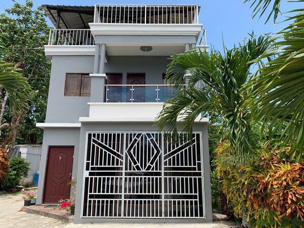 A two-story building with a gated entrance, a balcony on the upper floor, and a small garden area with plants and flowers. - Casa Linda: 10 Mins to Beach and lagoon with AC and Free Parking (Payita)