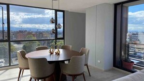 Un comedor moderno con una mesa redonda y seis sillas, ventanas amplias que ofrecen vista a la ciudad, y una lámpara colgante.