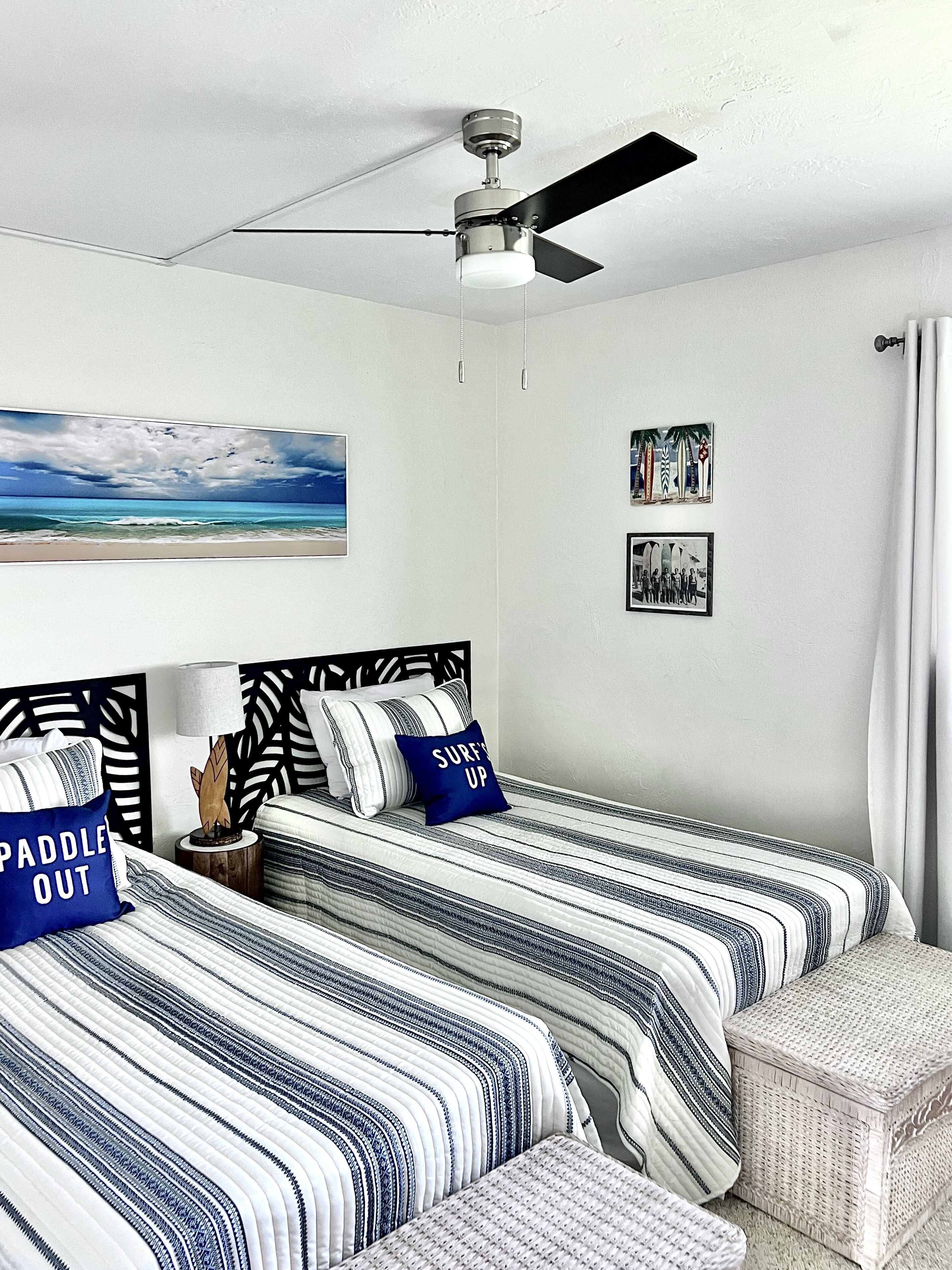 Een slaapkamer met twee bedden, een plafondventilator en een schilderij met een strandthema.