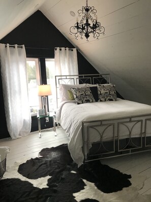Quarto com estrutura de cama em metal branco, tapete de couro bovino, lustre e janela com cortinas.