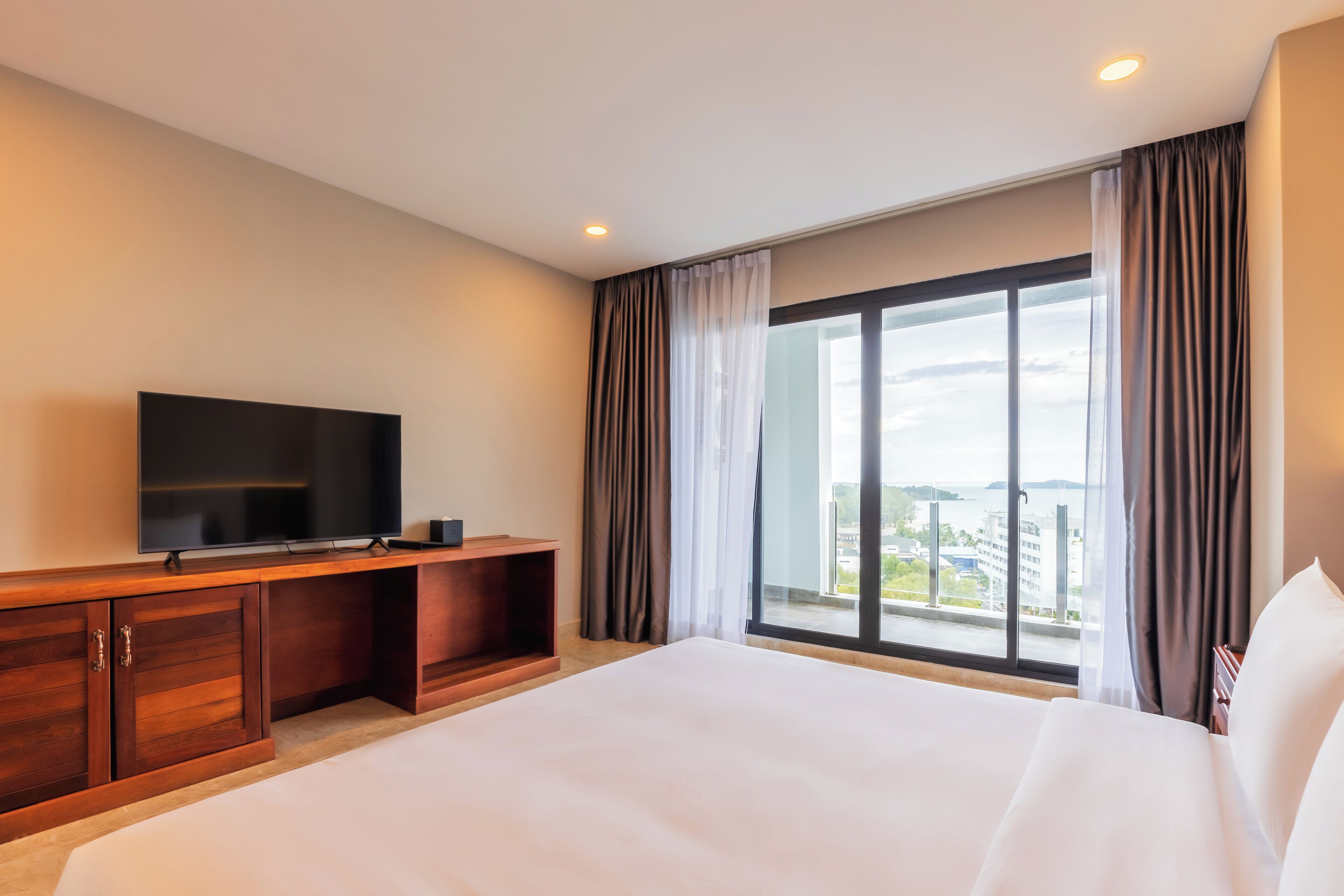 A hotel room with a large bed, a TV on a wooden console, and a balcony with curtains.