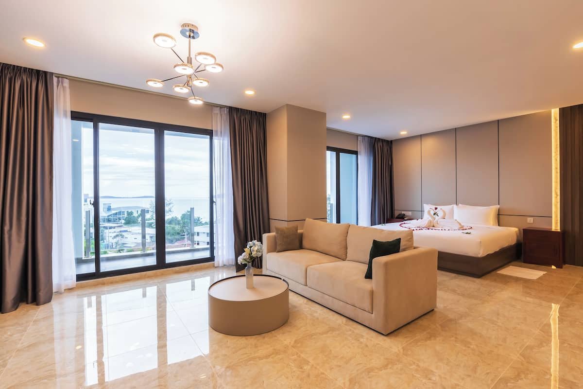 Une chambre d’hôtel moderne dotée d’un grand lit, d’un canapé, d’une table basse ronde et offrant une vue sur la ville grâce à de grandes fenêtres du sol au plafond.