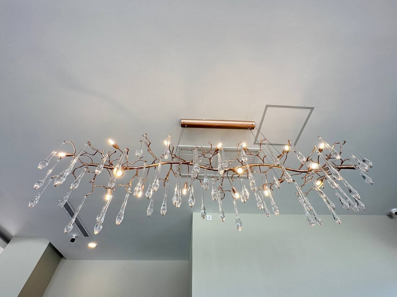 A modern chandelier with multiple branches and clear glass droplets, suspended from a ceiling in a minimalist room.