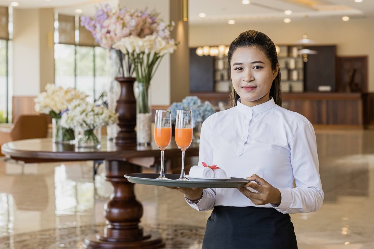 A hotel lobby with a reception desk, a round table with floral arrangements, and a waiter holding a tray with two glasses of orange juice and a small cake.