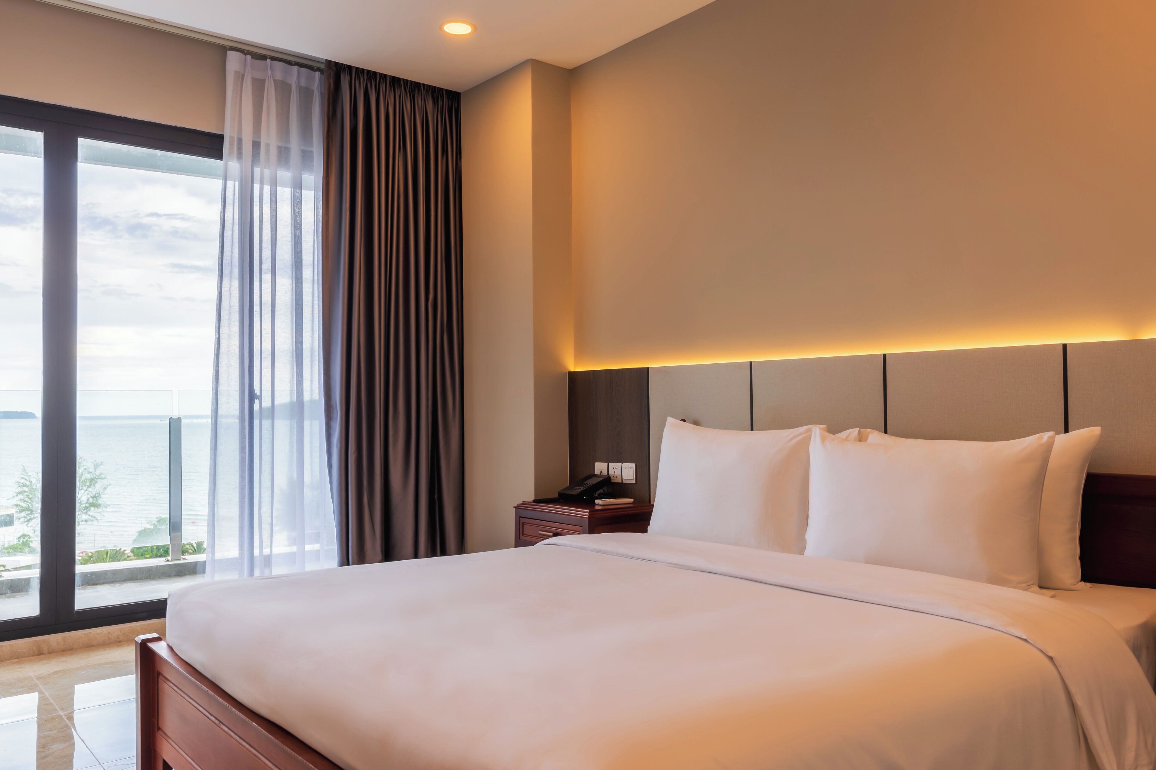 A hotel room with a large bed, a nightstand, and a view of the sea through a large window.