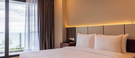 A hotel room with a large bed, a nightstand, and a view of the sea through a large window.