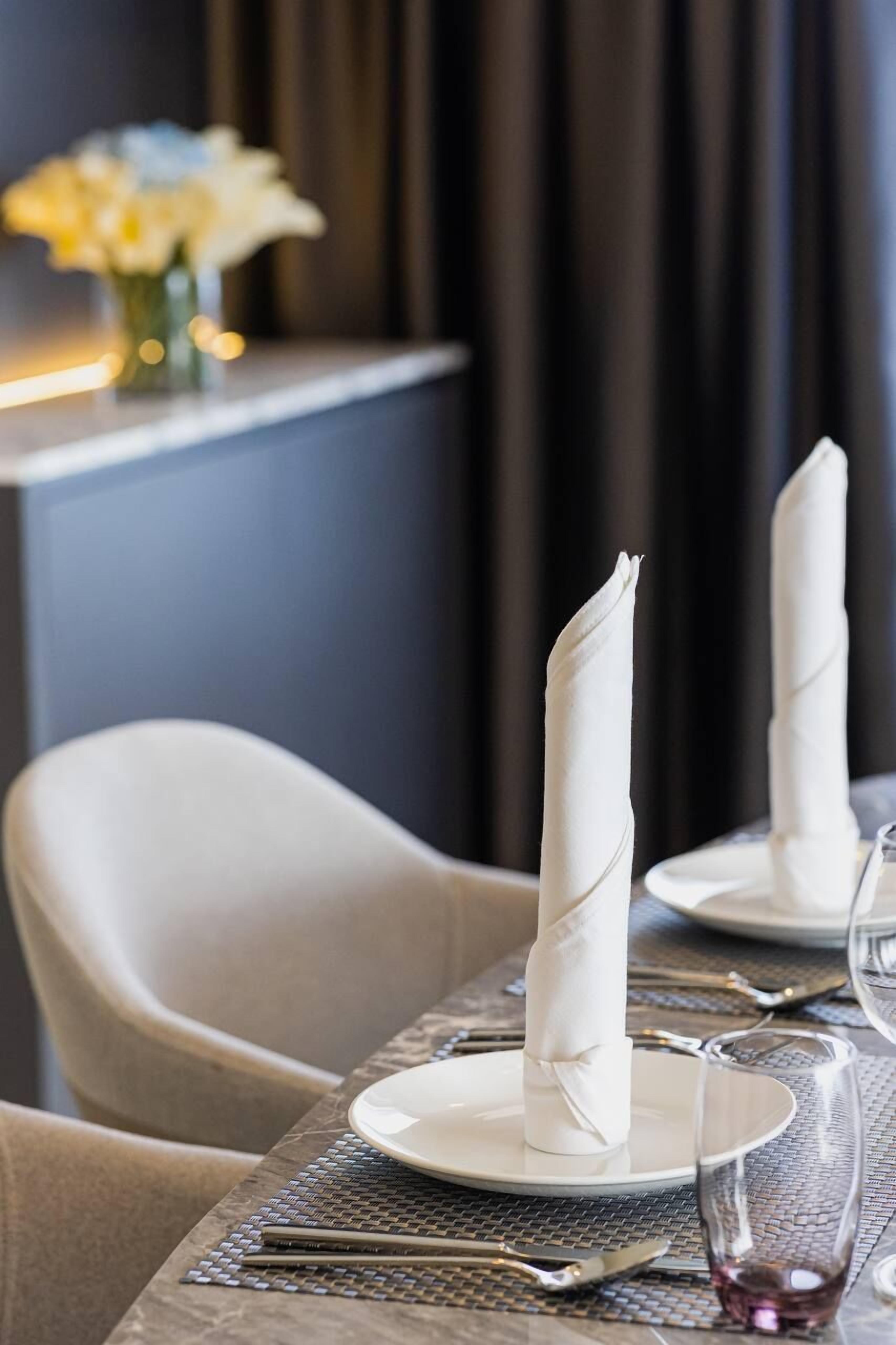 A dining table set with white napkins, plates, and cutlery, with a vase of flowers in the background.