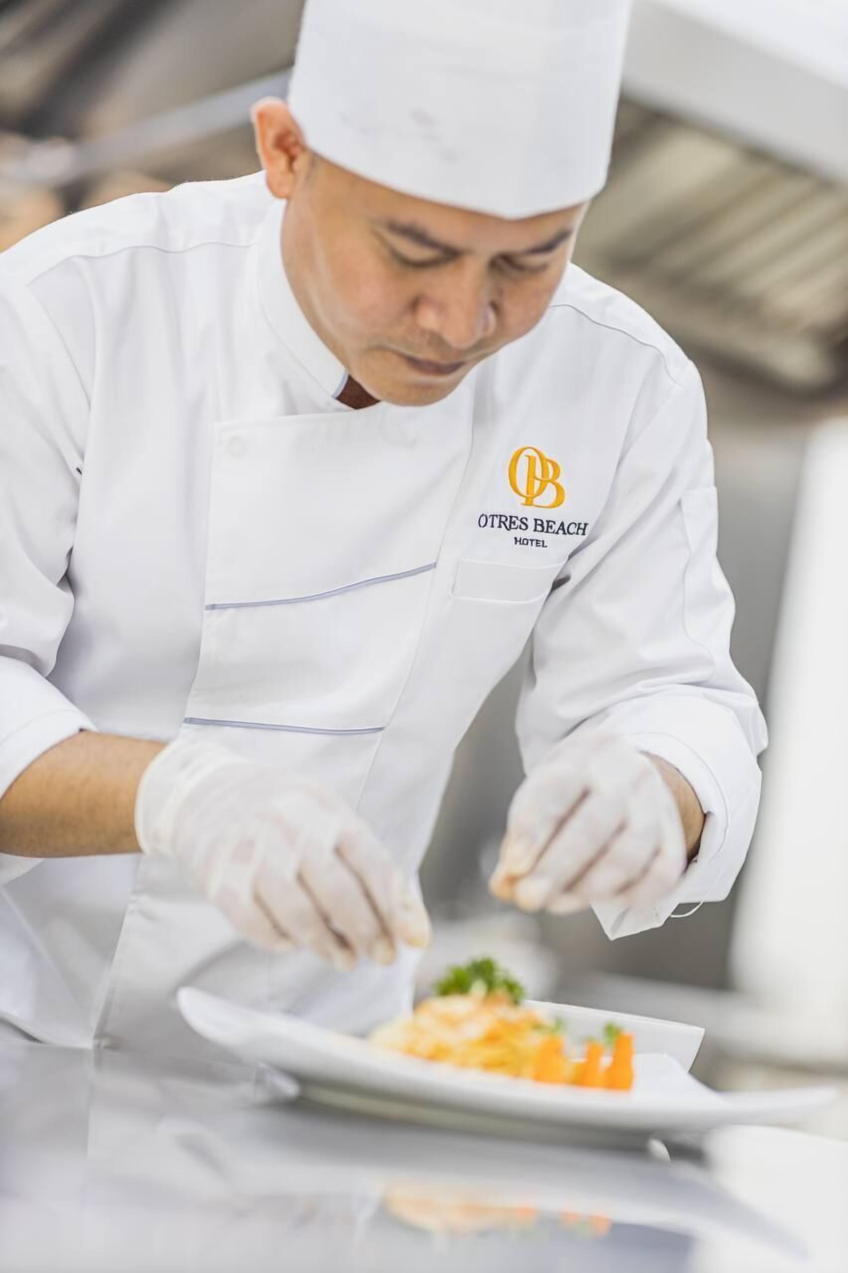 Un chef con uniforme blanco y un logotipo preparando comida sobre una mesa.