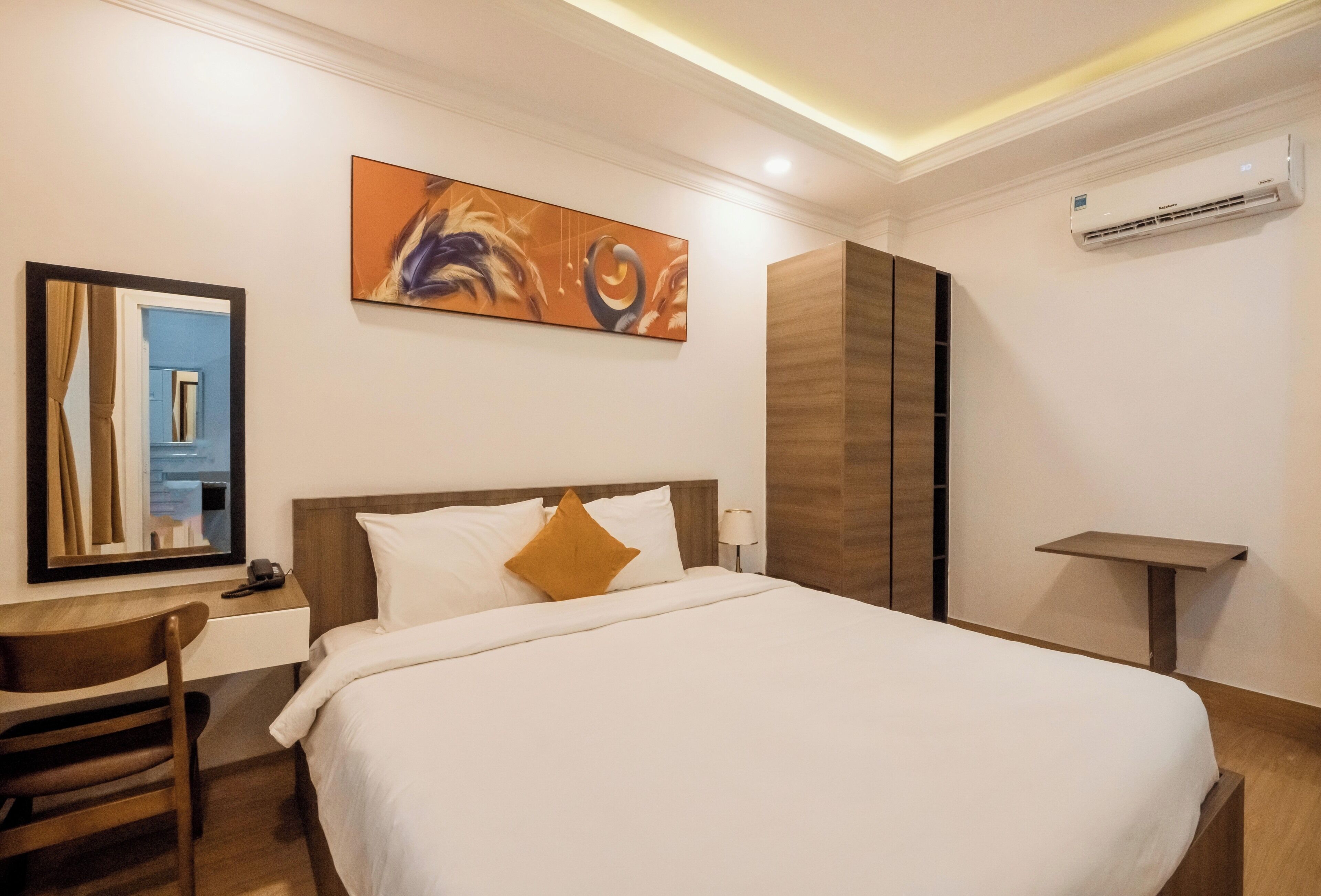 A hotel room with a bed, a desk, a chair, a painting, and an air conditioning unit.