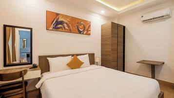 A hotel room with a bed, a desk, a chair, a painting, and an air conditioning unit.