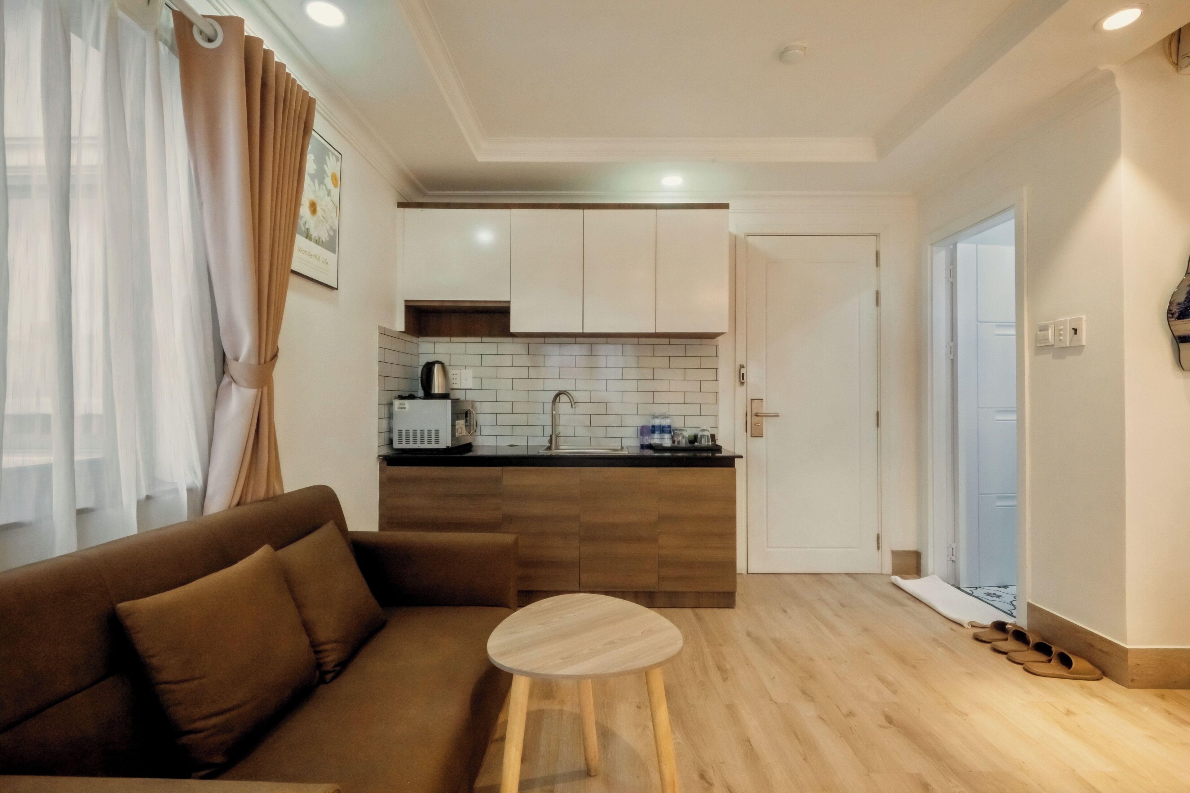 A modern living room with a sofa, a small wooden stool, a kitchen area with white cabinets, a tiled backsplash, and a door leading to another room.