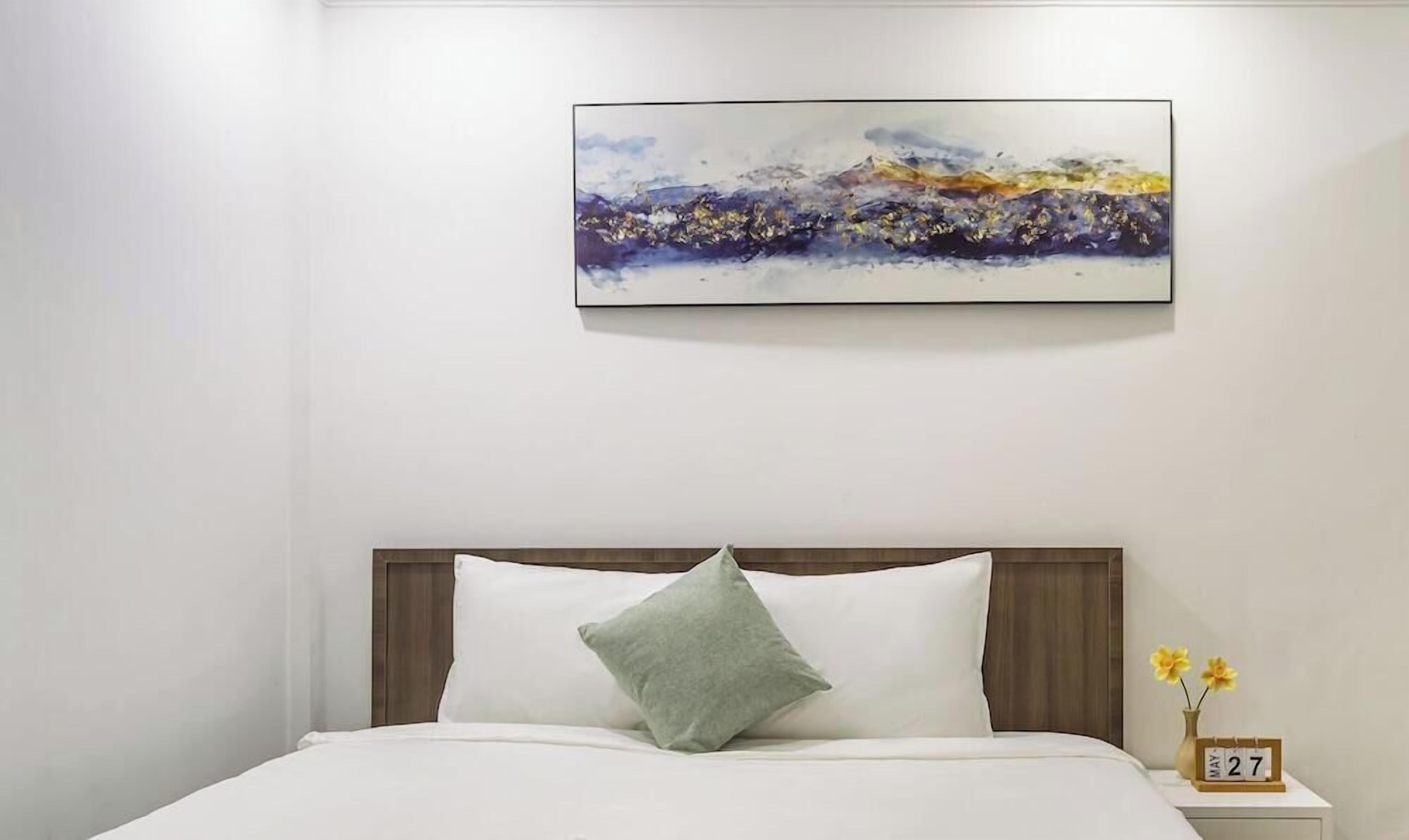A hotel room with a bed, a nightstand with a clock, and a painting on the wall.