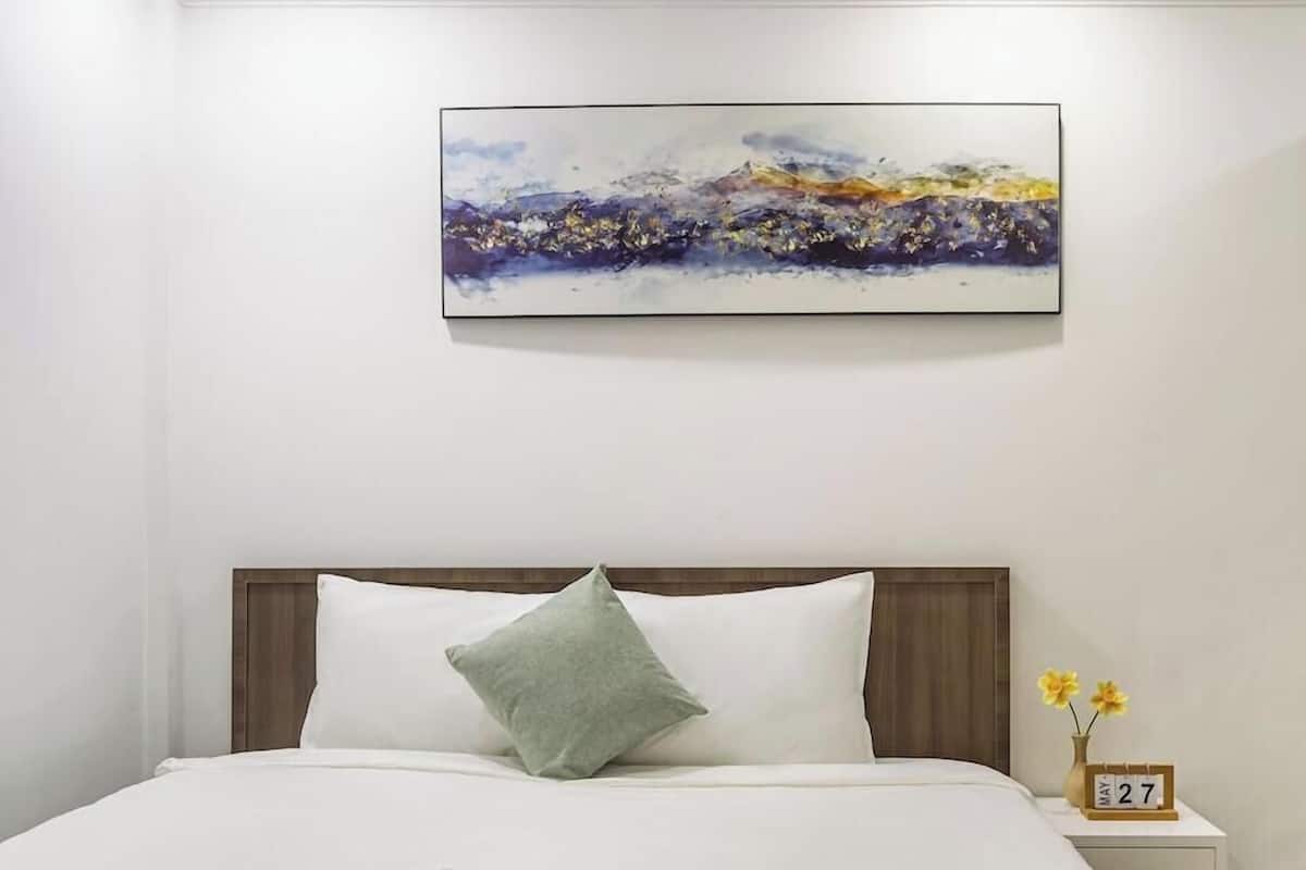 A hotel room with a bed, a nightstand with a clock, and a painting on the wall.